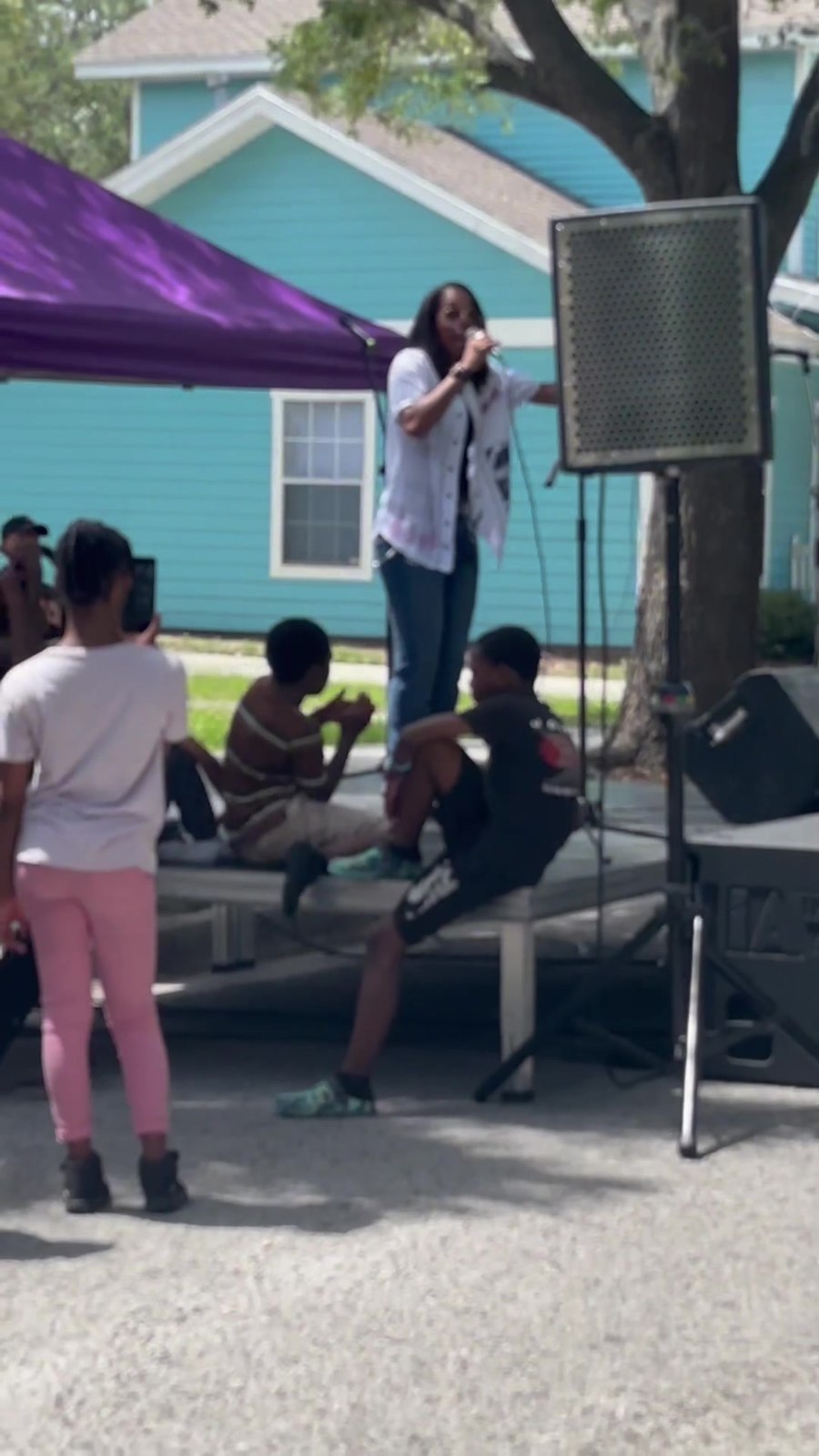A singer performs on a small outdoor stage while children watch from a nearby platform at a neighborhood concert