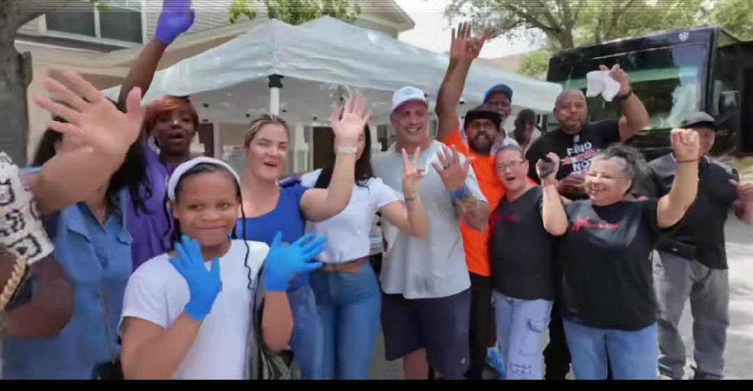 A large diverse group of neighbors and volunteers waving and cheering at a community outreach event