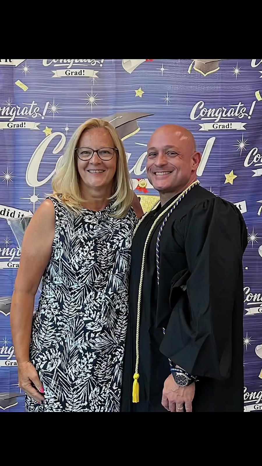 A trade school graduate in cap and gown poses with a family member in front of a Congrats Grad backdrop